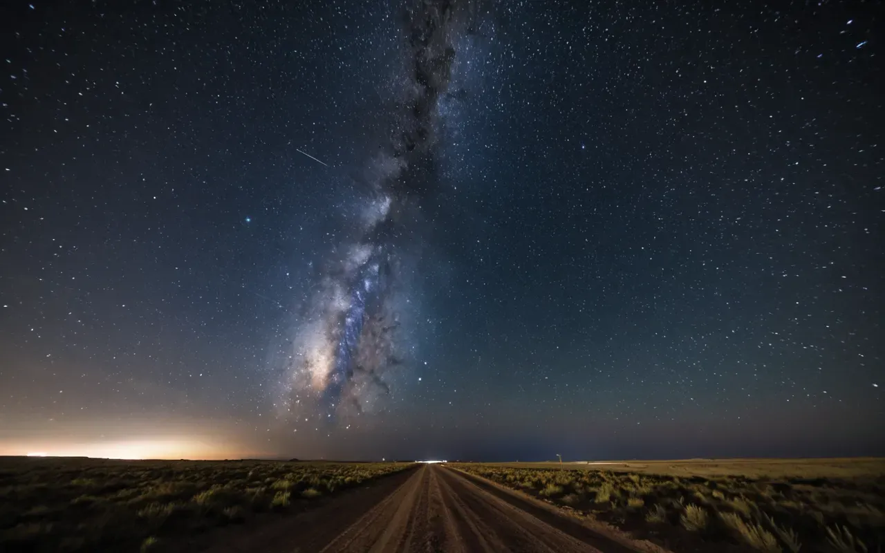 A wide angle shot of a clear night sky over a flat landscape, showing the Milky Way as a bright, dense band of stars stretching diagonally across the frame, with a large, smooth stone pillar silhouetted in the foreground against the cosmic light.